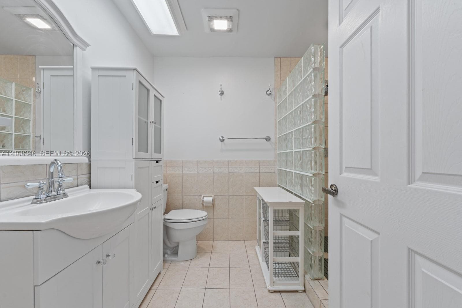 Bathroom featuring a white vanity cabinet with an integrated sink, ceramic tile flooring, neutral wall-to-wall ceramic tiling, and a glass block privacy partition