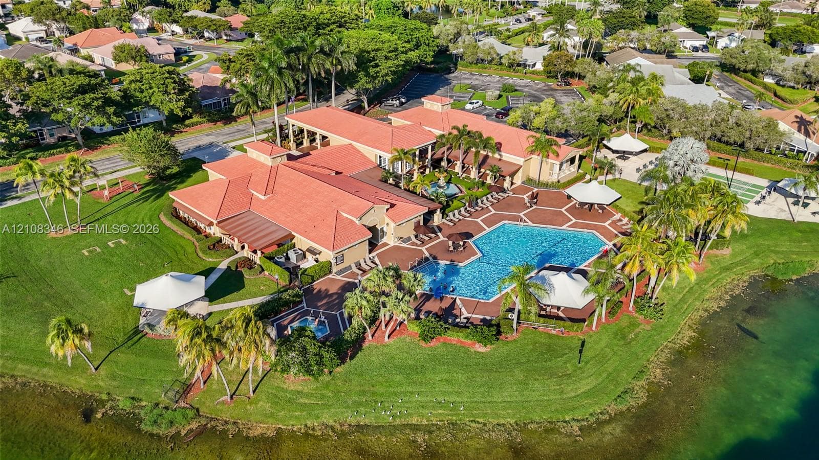 Aerial view of a clubhouse in the community center featuring a tile-roofed structure, a large swimming pool, an attached spa, paved patio decking, and landscaped grounds