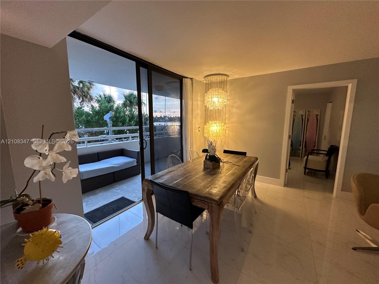 Dining area featuring large-format light-toned flooring, tiered crystal chandelier, and floor-to-ceiling glass sliding doors opening to a covered balcony