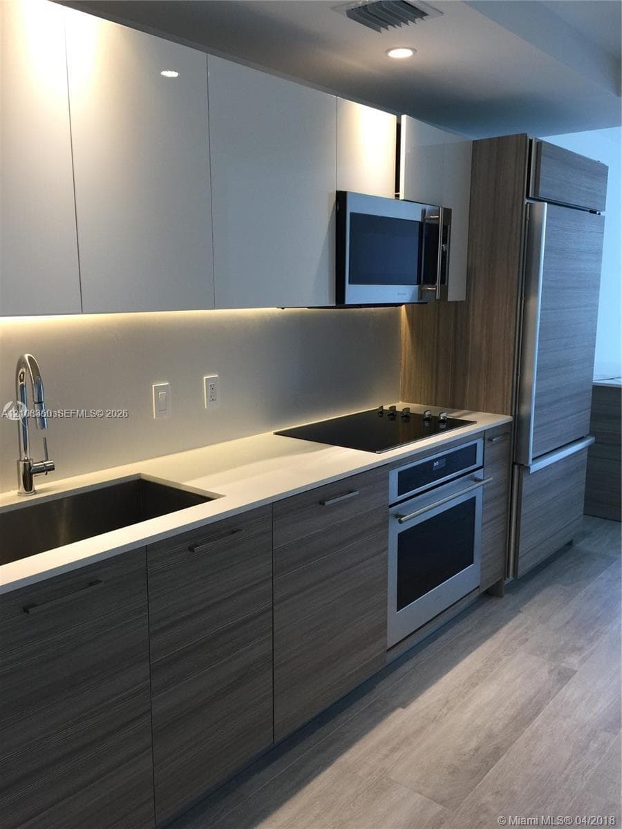 Kitchen featuring wood-finish lower cabinetry, white gloss upper cabinetry, stone countertops, stainless steel appliances, and integrated under-cabinet lighting
