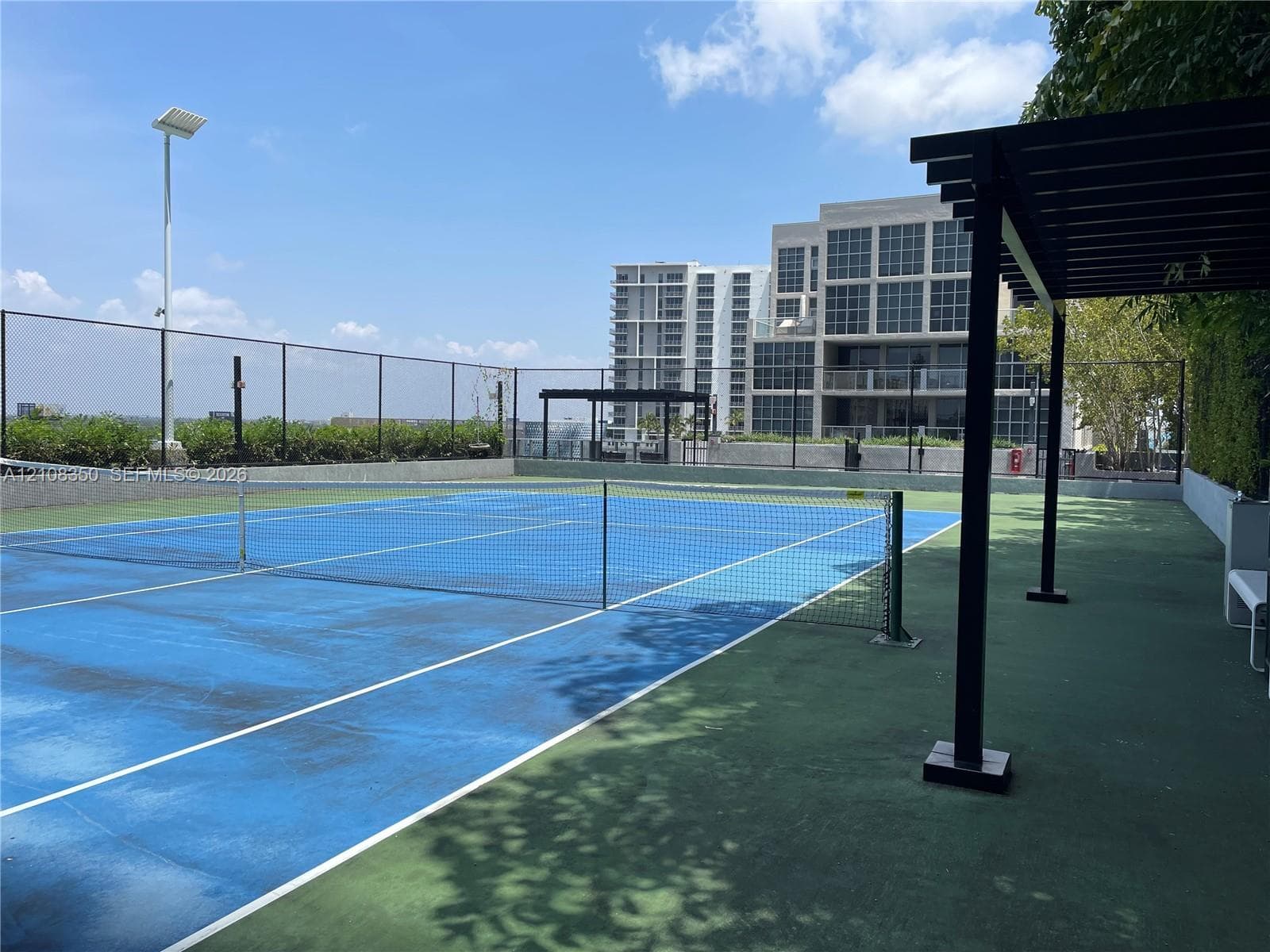 Rooftop tennis court featuring a perimeter fence, overhead lighting, and a metal pergola structure