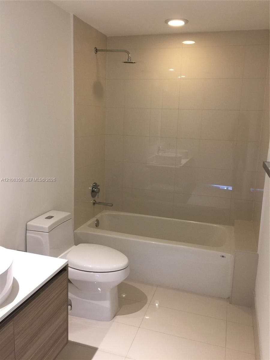 Full-height neutral wall tiling, bathtub with integrated shower, vanity with wood-finish cabinetry, white countertop, and recessed ceiling lighting