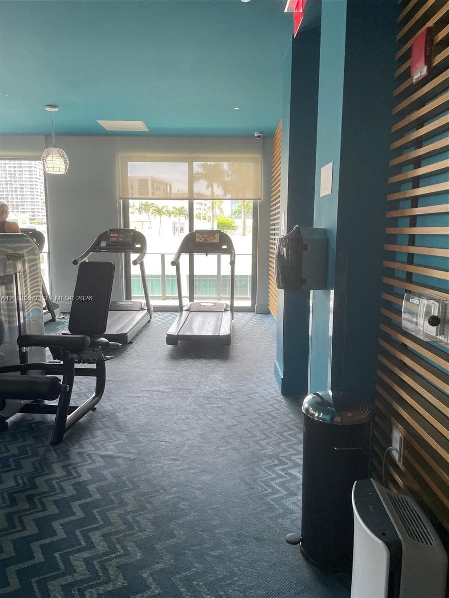 Fitness center featuring chevron-patterned carpeting, decorative wood-slat wall paneling, and floor-to-ceiling glass sliding doors with solar shades