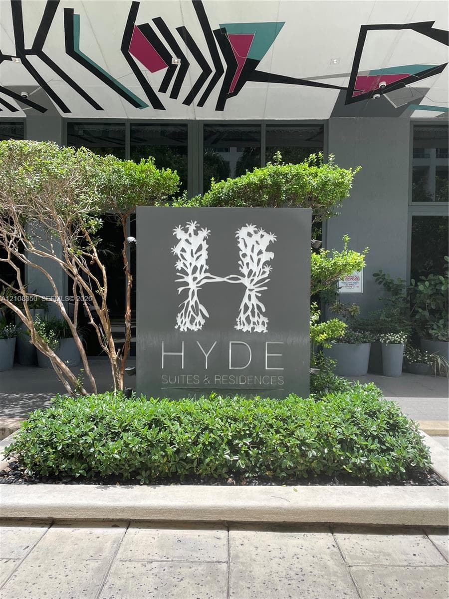 Property entrance featuring custom signage, concrete masonry, manicured shrubbery, mature landscaping, and structural ceiling art