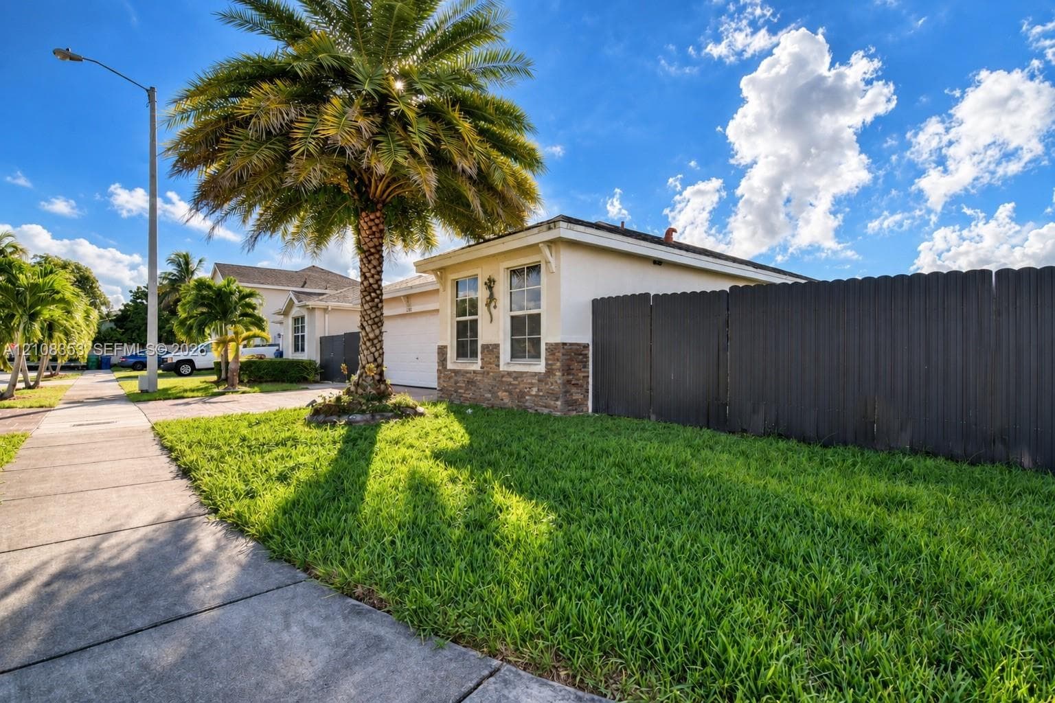 Exterior featuring a stucco facade with stone veneer accents, multi-pane windows, a dark privacy fence, and a palm tree within a grass lawn