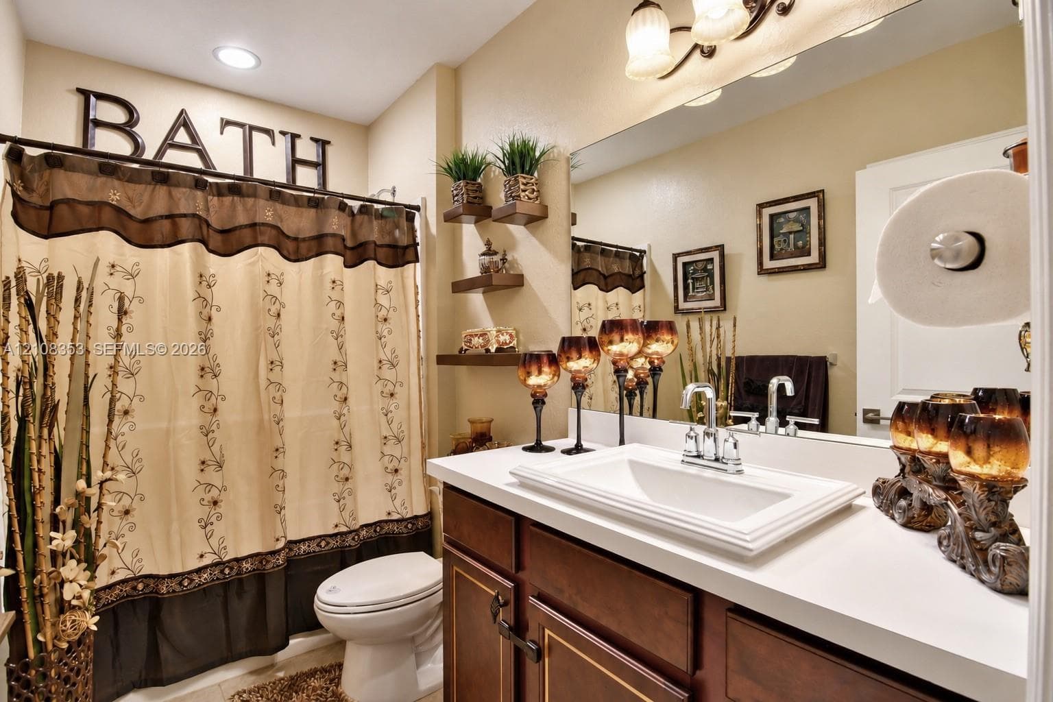 Bathroom featuring a dark wood vanity with a light-toned countertop, an integrated rectangular sink, wall-mounted tiered shelving, and a full-width vanity mirror with integrated lighting