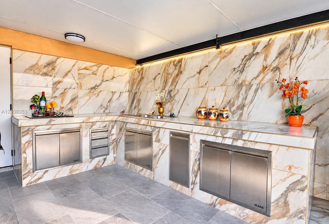 Outdoor kitchen featuring large-format natural stone cladding, stainless steel cabinetry, integrated cooktop, built-in sink, and LED accent lighting