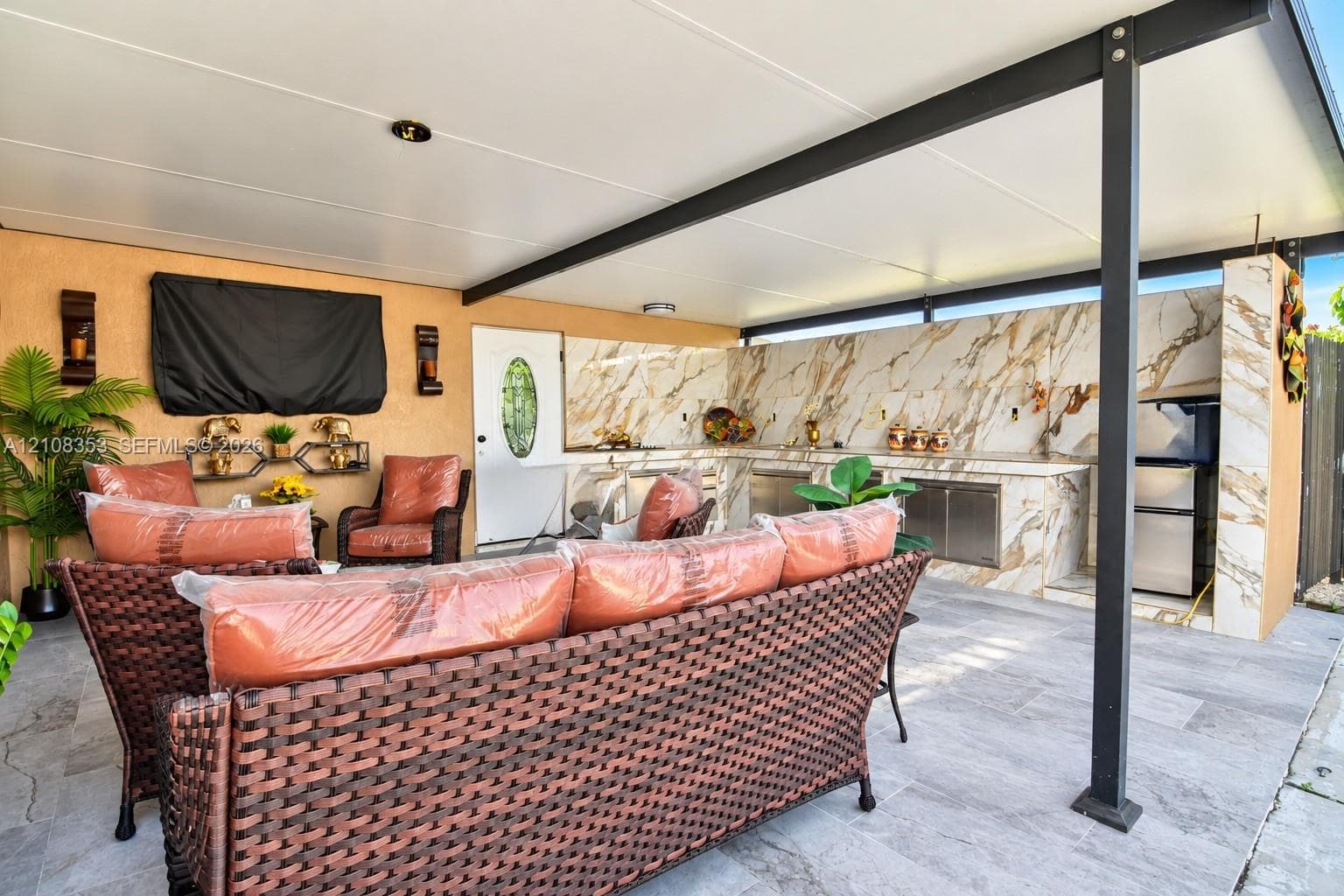 Covered outdoor patio featuring large-format tile flooring, a built-in outdoor kitchen with stone-patterned backsplash, stainless steel appliances, and a structural metal support frame