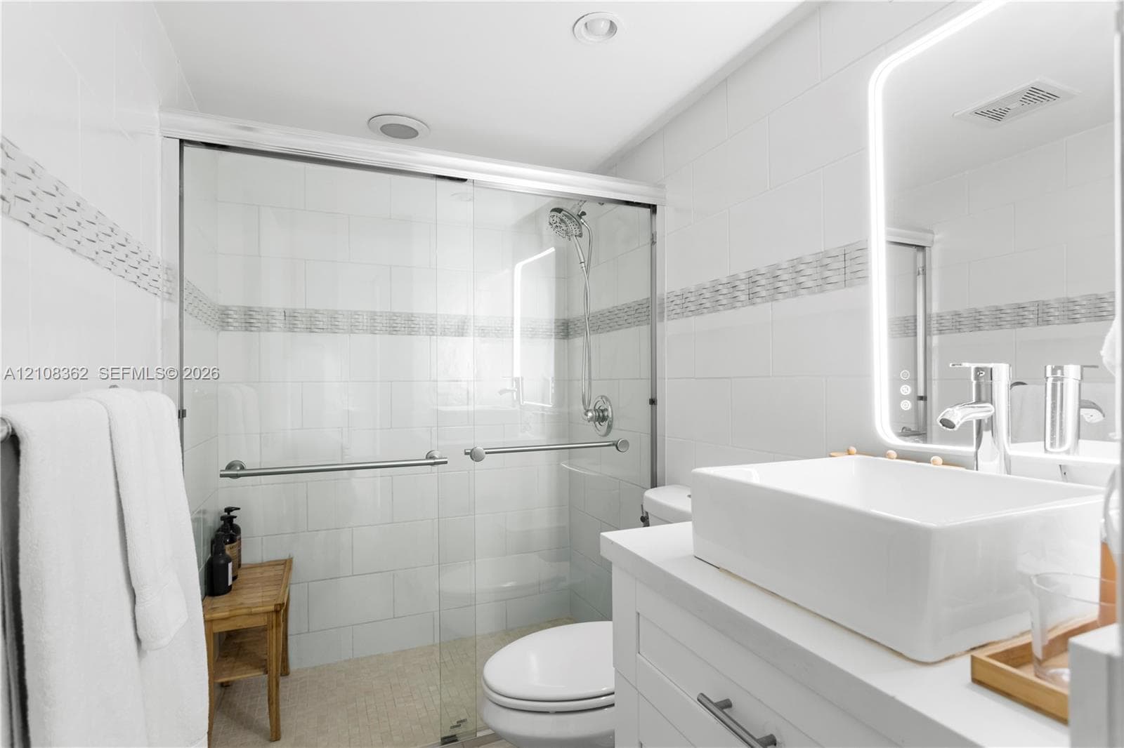 Walk-in shower with sliding glass doors, white subway-style wall tiling, decorative mosaic tile inlay, modern vessel sink with chrome faucet, and integrated LED backlit mirror