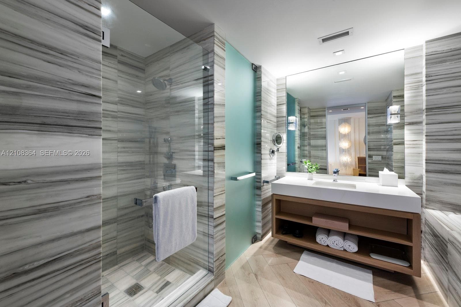 Bathroom featuring floor-to-ceiling stone-tiled walls, a walk-in shower with a glass enclosure, a floating vanity with open shelving, a large wall-mounted mirror, and wood-finish flooring in a herringbone pattern