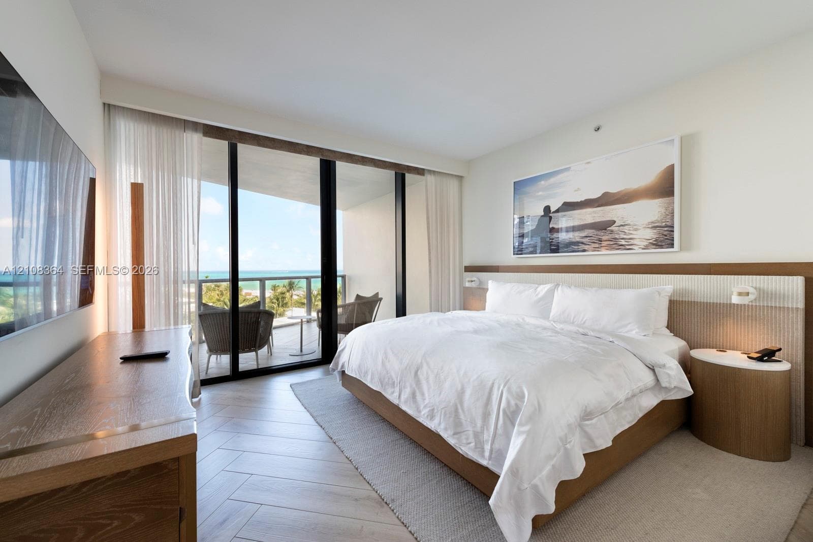 Floor-to-ceiling sliding glass doors, private balcony, wood-finish flooring in a herringbone pattern, and built-in headboard with integrated lighting