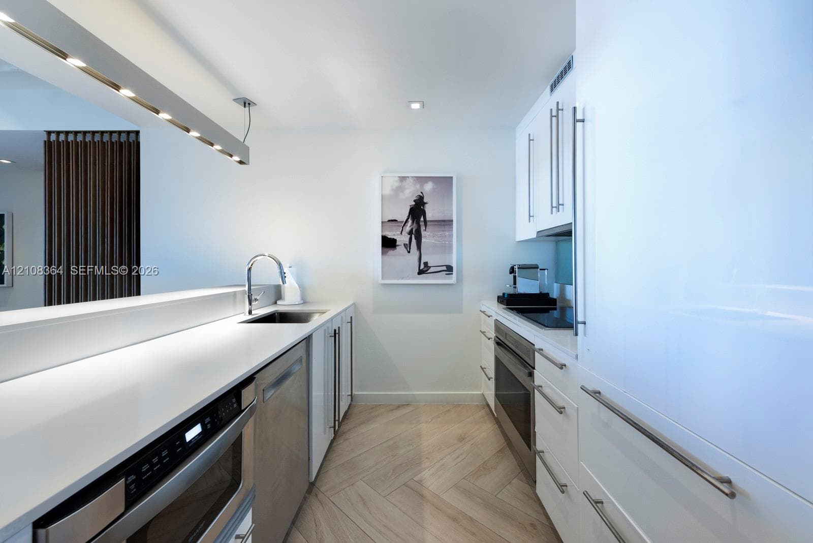 Galley kitchen featuring white cabinetry, solid surface countertops, stainless steel appliances, and herringbone-patterned wood-finish flooring