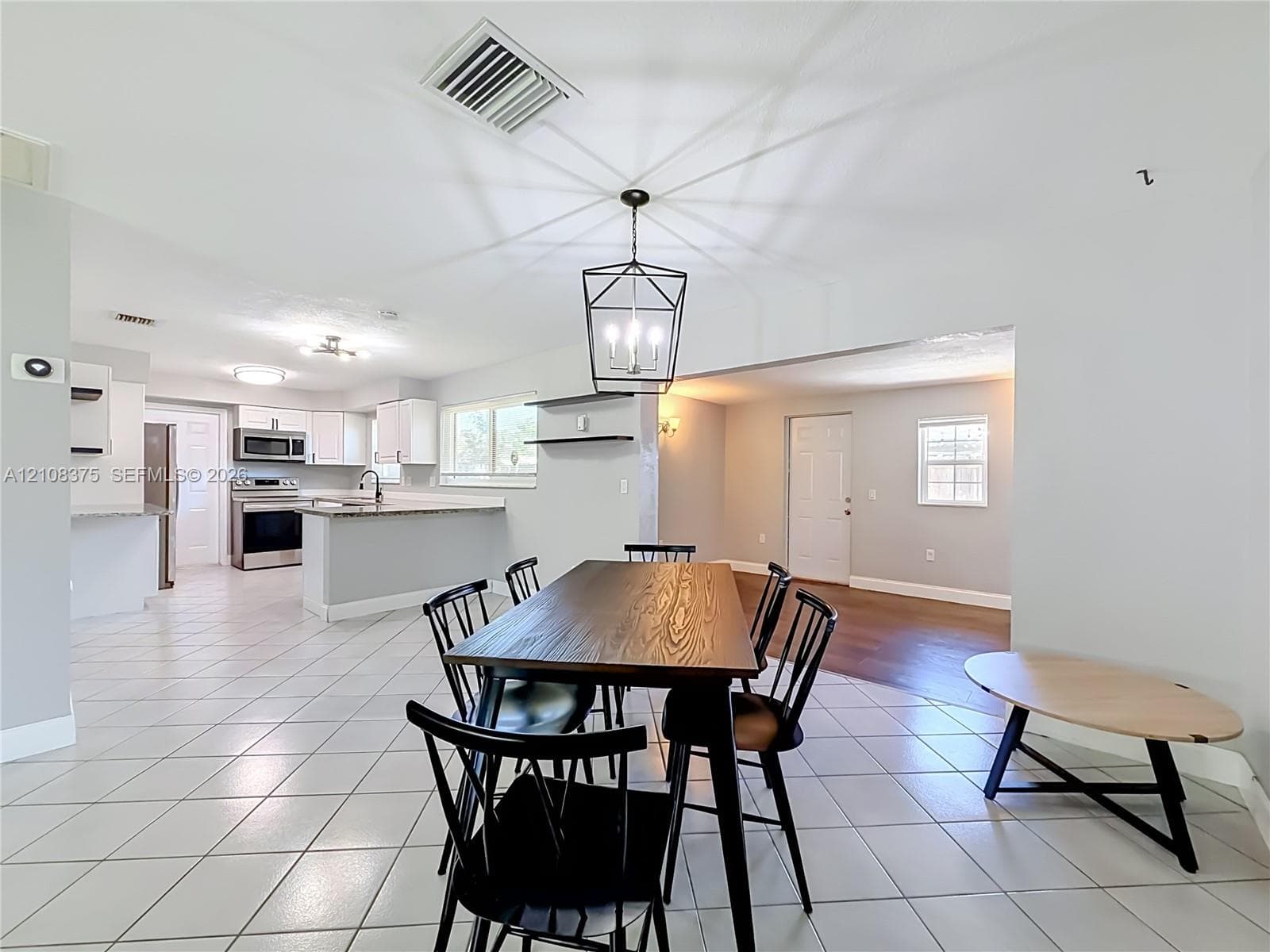 Open-concept floor plan featuring ceramic tile flooring, kitchen with white cabinetry, stone-finish countertops, stainless steel appliances, and a geometric metal pendant light fixture