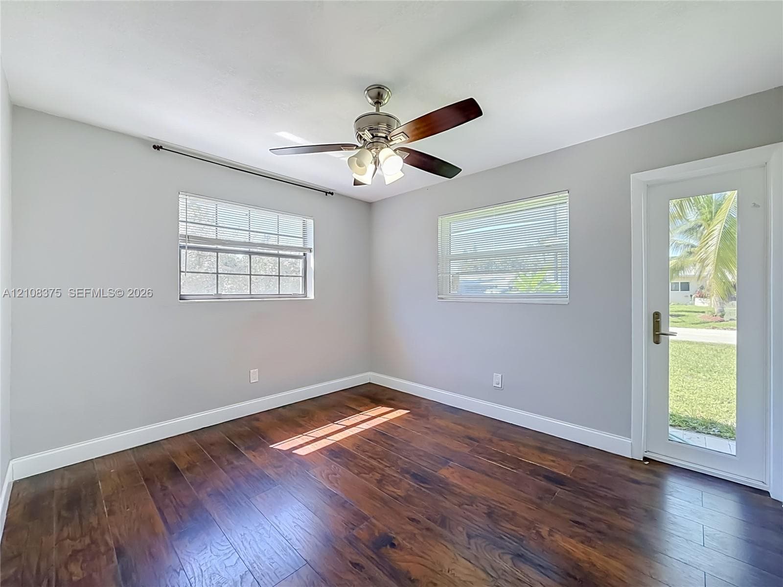 Room featuring wood-finish flooring, a multi-blade ceiling fan with integrated lighting, dual windows with horizontal blinds, and a glass-paneled exterior door