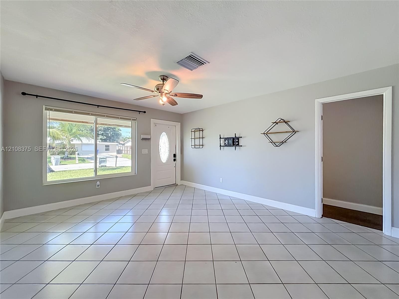 Neutral-toned interior featuring large-format ceramic tile flooring, a multi-pane window with hardware, a solid-panel front door with an oval glass insert, and a ceiling-mounted fan with integrated lighting
