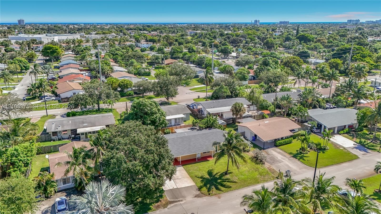 Aerial perspective of a single-story residence featuring a shingled gable roof, concrete driveway, and expansive lawn with mature palm trees