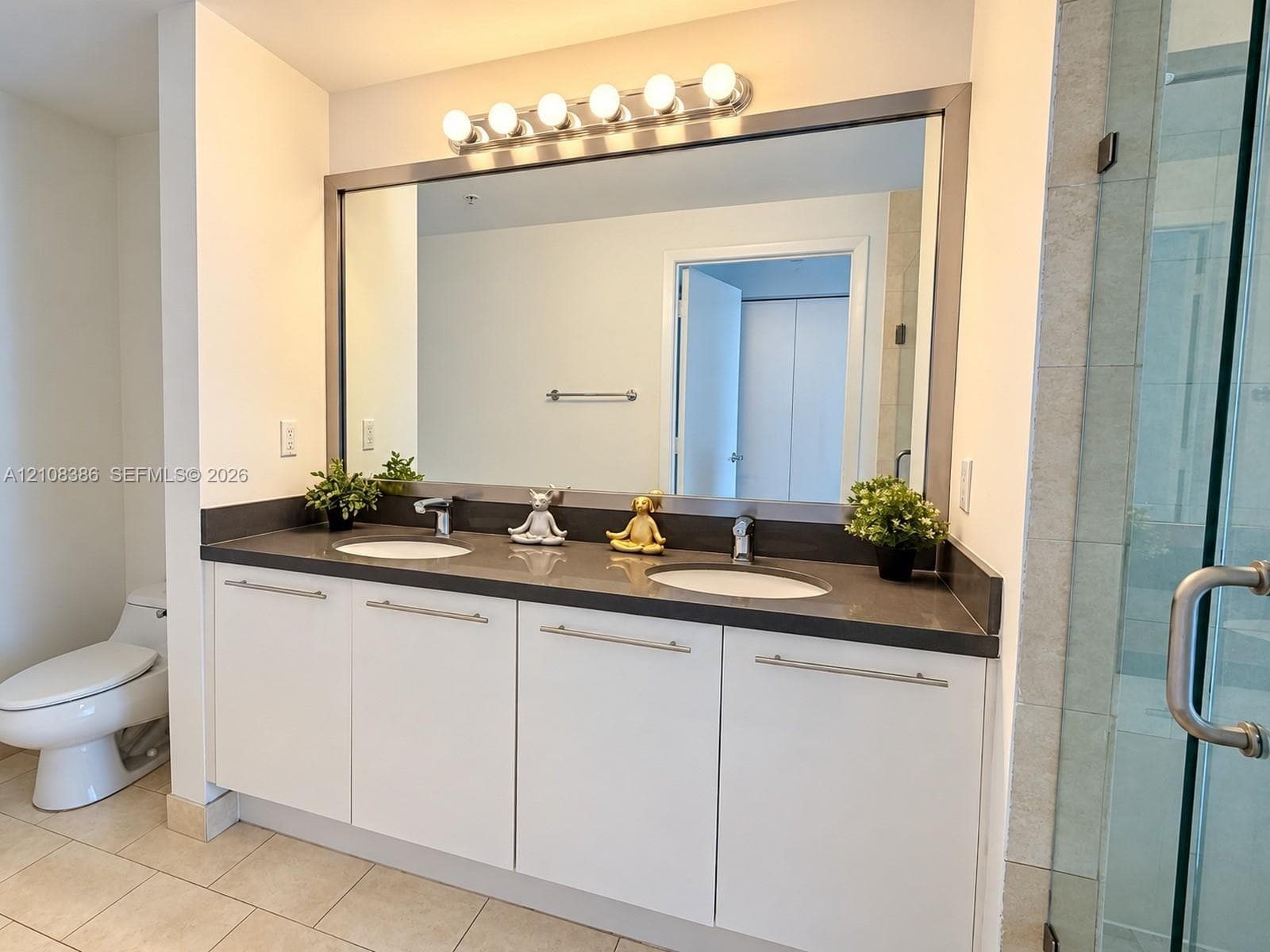 Dual-vanity cabinet with white flat-panel cabinetry, dark stone-composite countertop, undermount sinks, and a large-format metallic-framed mirror with multi-bulb vanity lighting