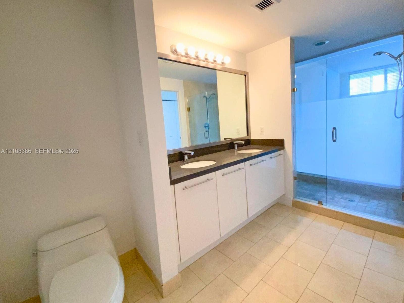Bathroom featuring a dual-sink vanity with white cabinetry, dark stone-composite countertop, full-width mirror, and a glass-enclosed walk-in shower with tile flooring