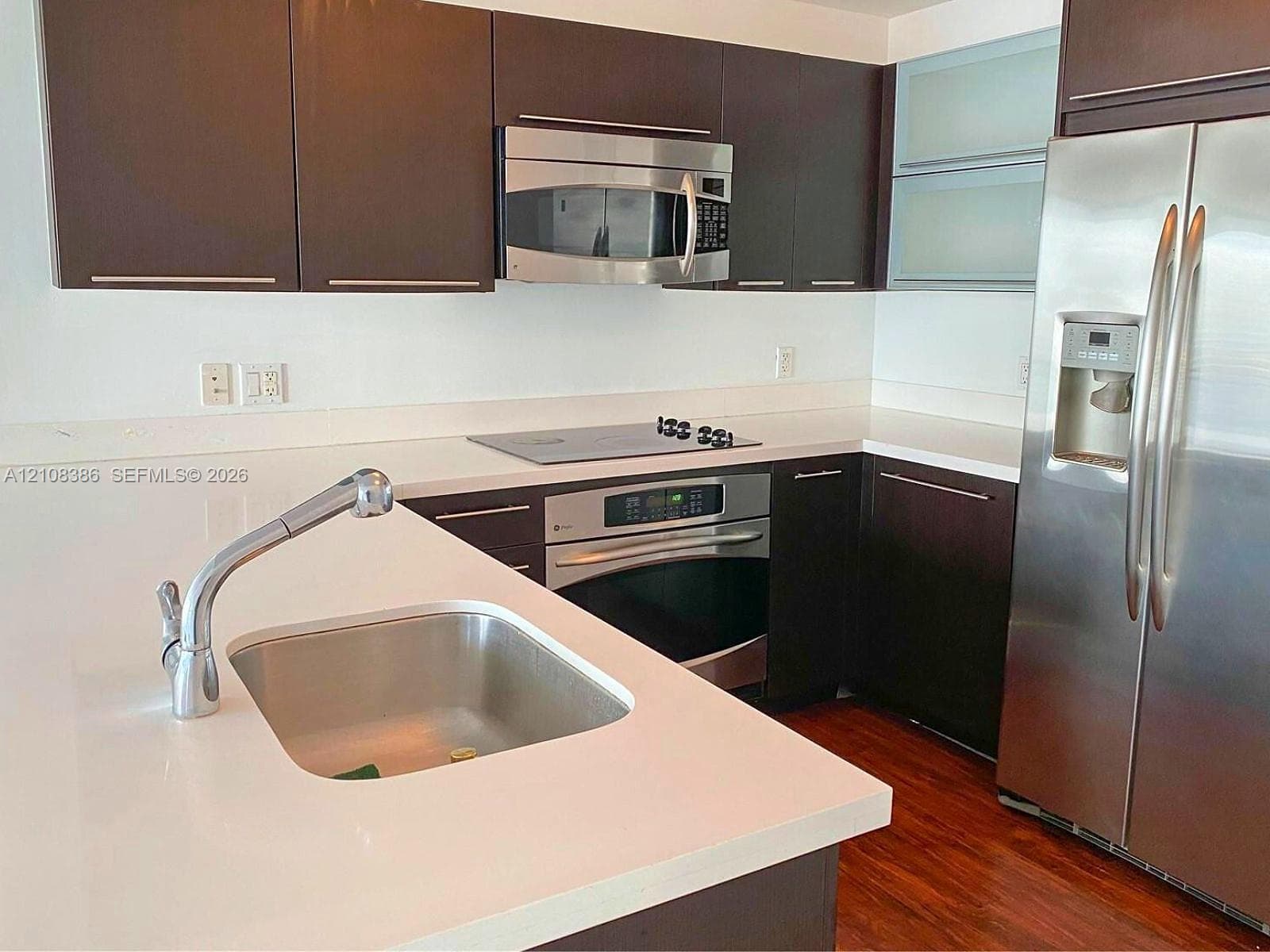Kitchen featuring dark cabinetry, solid-surface countertops, wood-finish flooring, and stainless steel appliances including a built-in oven, microwave, and refrigerator
