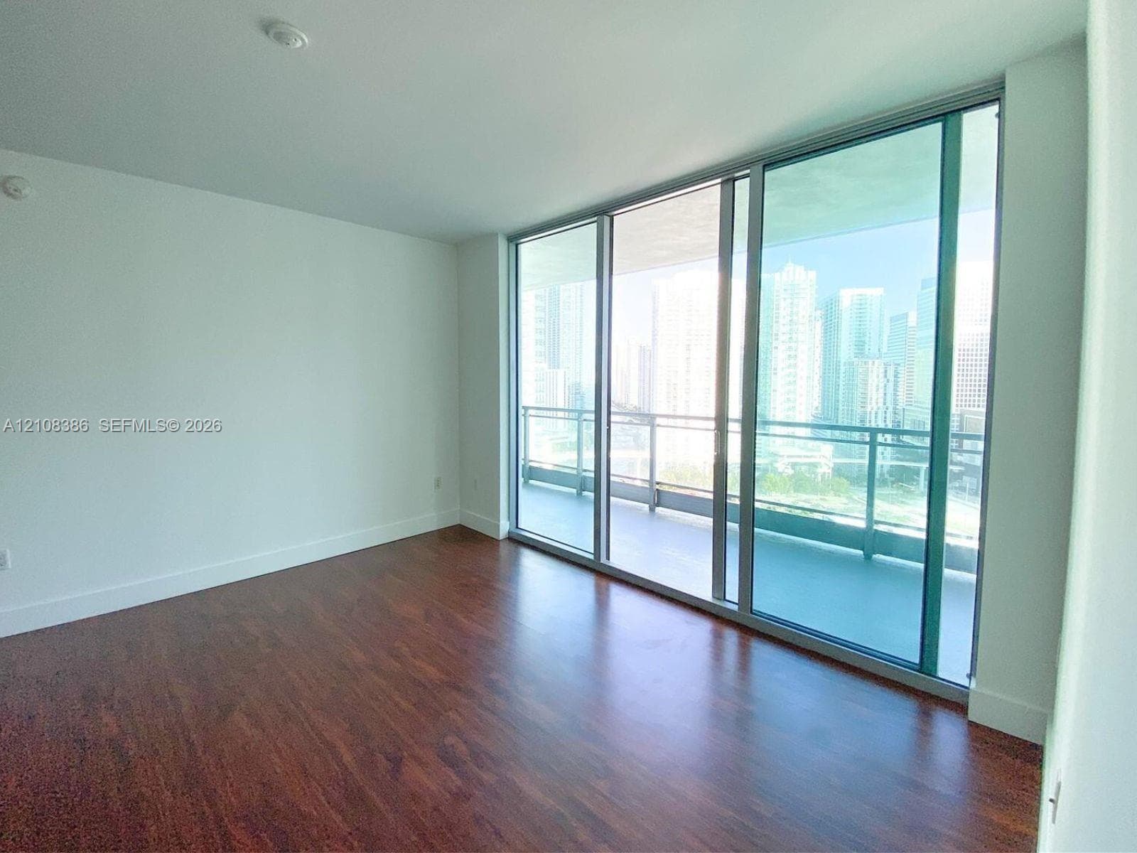 Floor-to-ceiling glass sliding doors, private balcony, wood-finish flooring, and expansive urban skyline backdrop