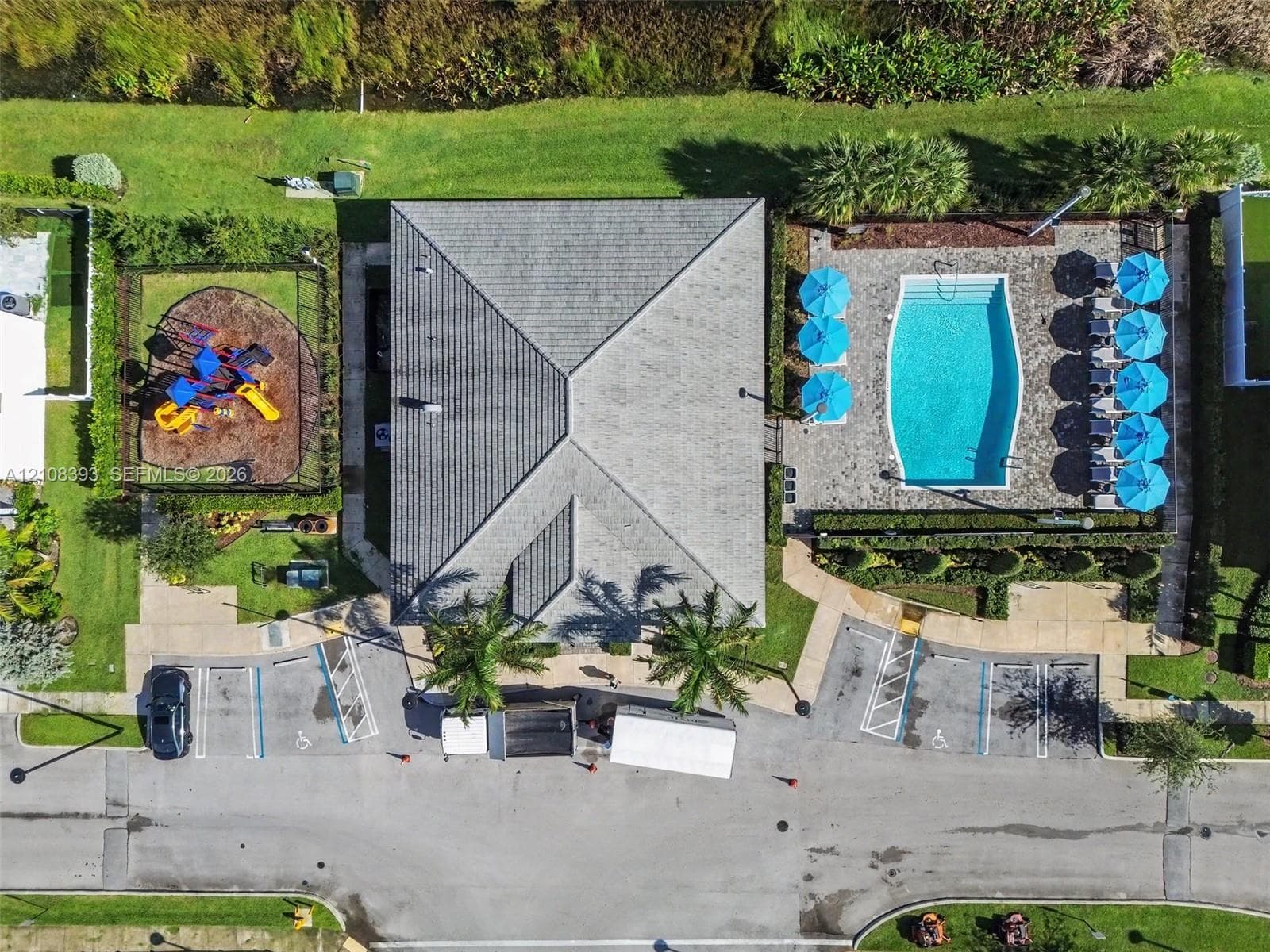 Aerial perspective of a community facility featuring a swimming pool, paved pool deck, enclosed recreation area, and paved parking area