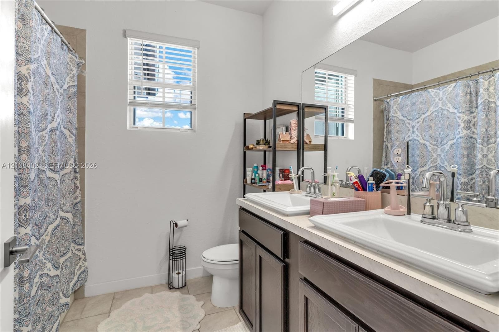 Dual-sink vanity with dark cabinetry, light-toned countertop, and chrome fixtures, complemented by tile flooring and large wall-mounted mirror