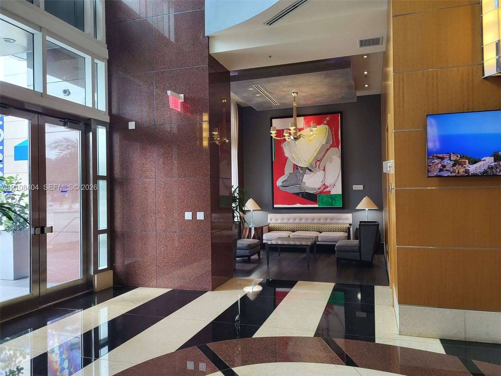 Lobby featuring high-polish stone flooring in a geometric pattern, polished red stone accent wall, wood-paneled wall surfaces, multi-tiered metal chandelier, and floor-to-ceiling glass entrance doors