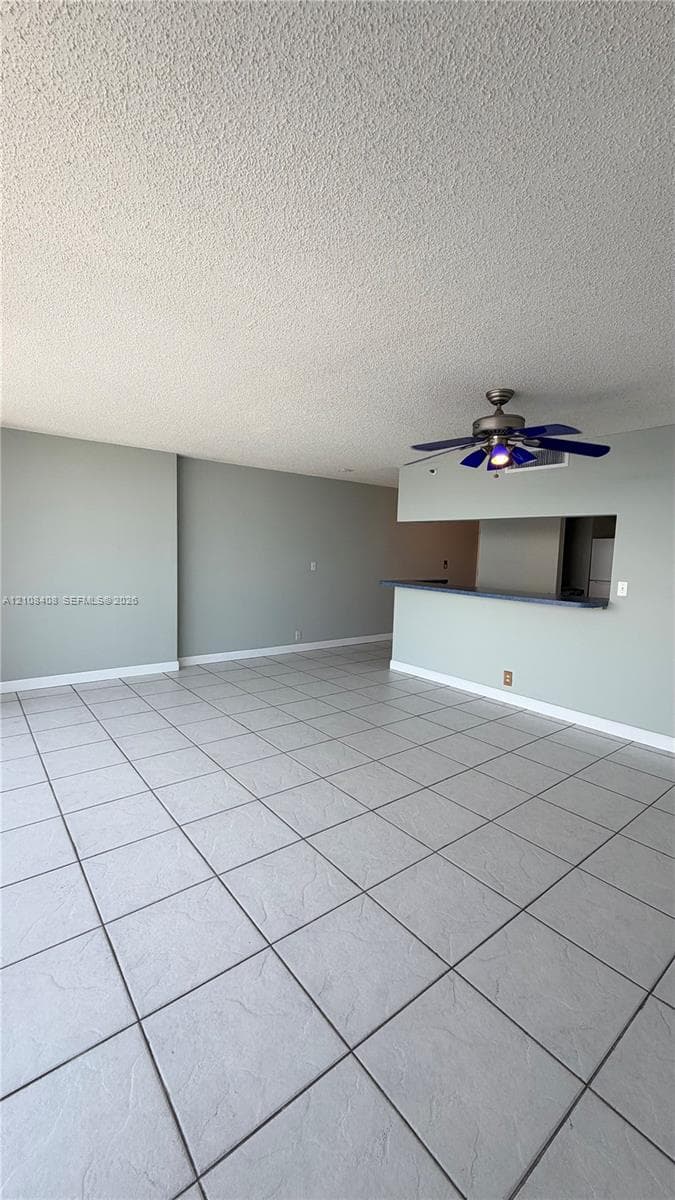 Large-format ceramic tile flooring, neutral wall tones, and a peninsula-style breakfast bar featuring a pass-through counter and a multi-blade ceiling fan with integrated lighting