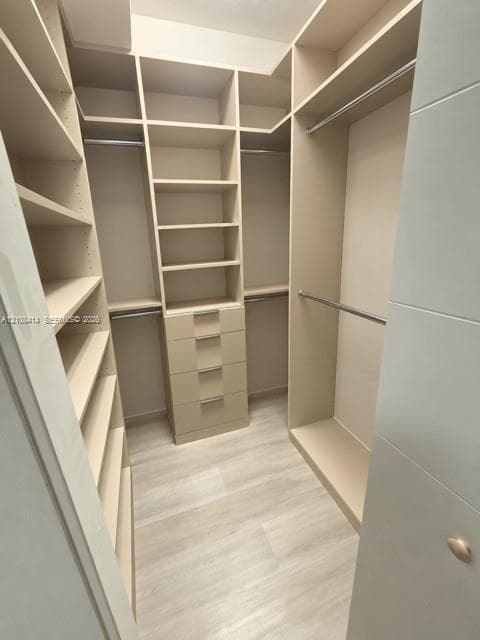 Walk-in closet featuring extensive custom built-in shelving, four integrated storage drawers with brushed metallic pulls, tiered hanging rods, and light-toned wood-finish flooring