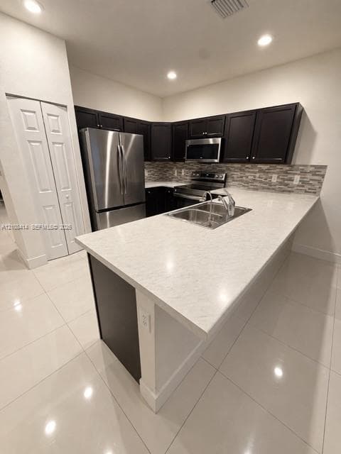 Kitchen featuring a stone-surface island, dark-toned cabinetry, stainless steel appliances, multi-tonal backsplash, and tile flooring