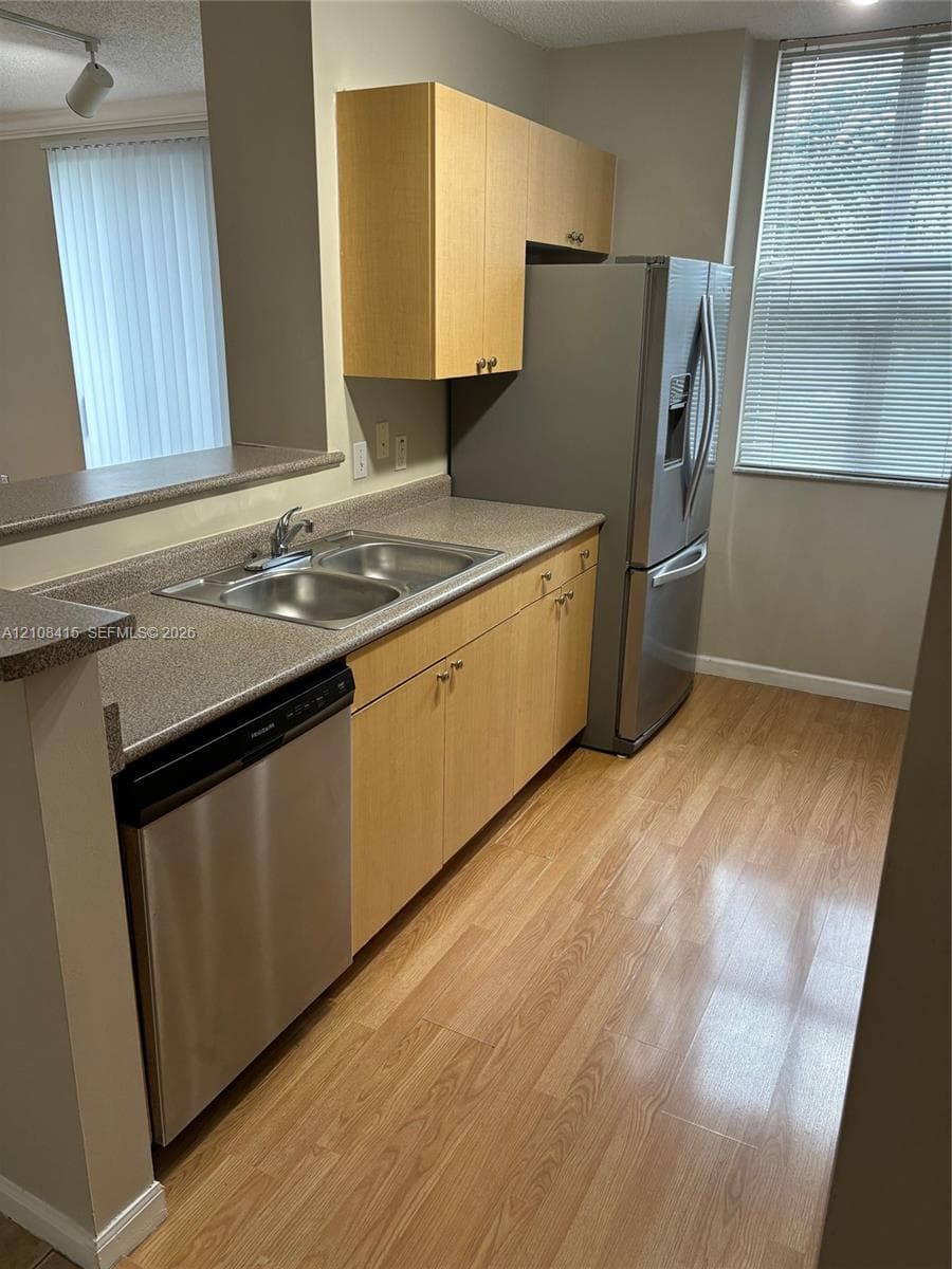 Kitchen featuring light wood-finish cabinetry, stainless steel appliances, speckled solid-surface countertops, a double-basin stainless steel sink, and wood-finish flooring