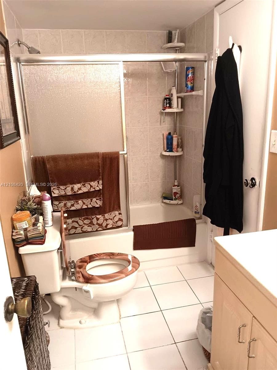 Bathroom featuring a tub-shower combination with a sliding glass door enclosure and light-toned tile surround, white square floor tiling, and wood-finish cabinetry