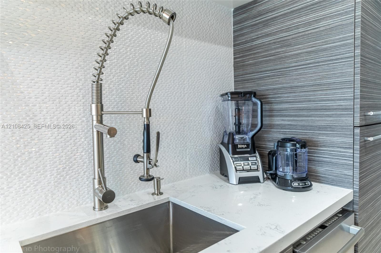 Stainless steel undermount sink with a professional-style gooseneck pull-down faucet, white stone-composite countertop, textured white tile backsplash, and horizontal-grain cabinetry