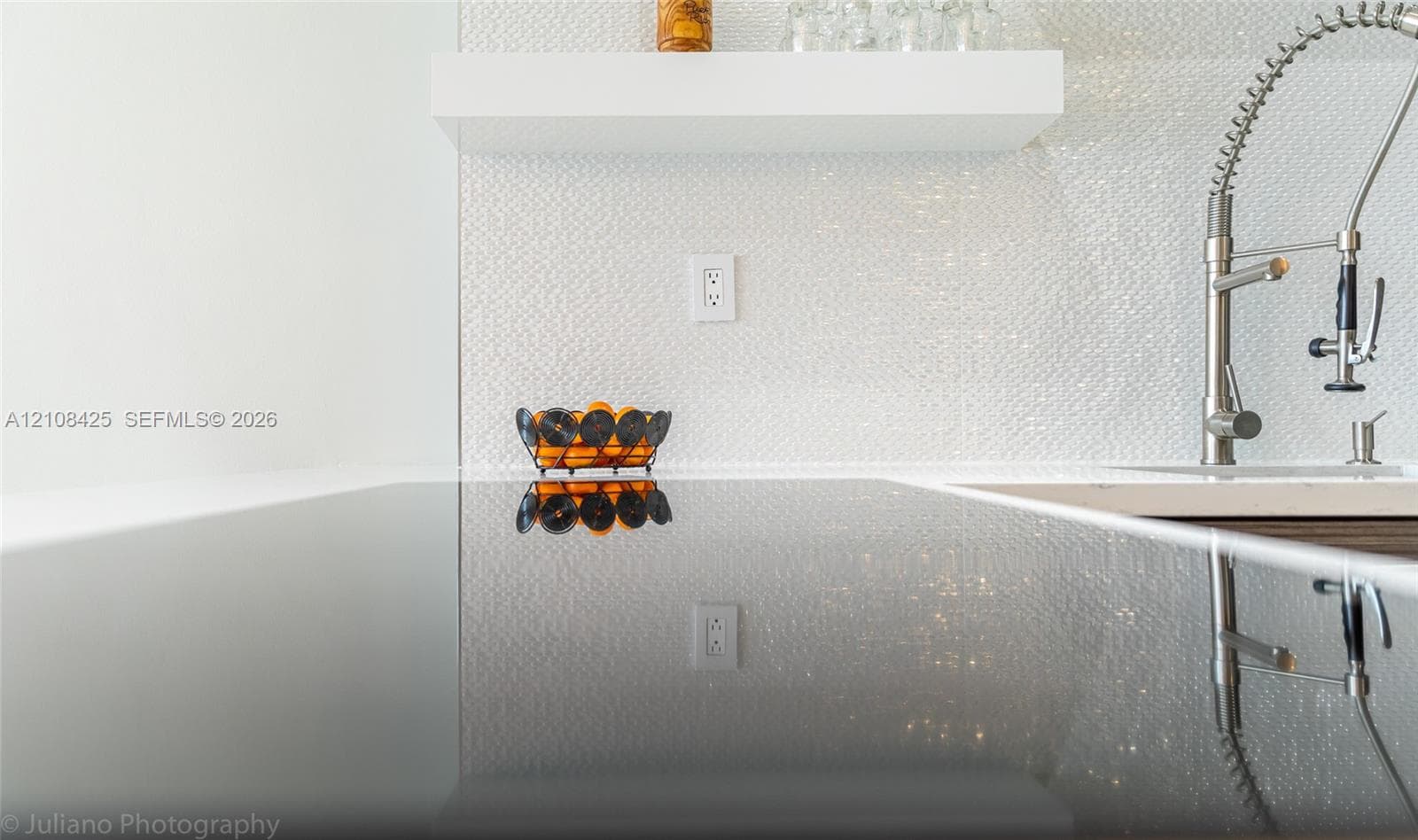 Polished solid-surface countertop with an integrated cooktop, textured white tile backsplash, built-in floating shelf, and stainless steel high-arc pull-down faucet