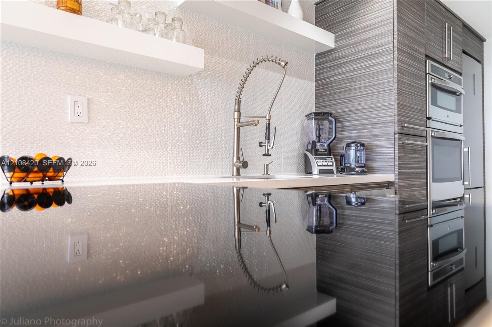 Textured white backsplash, polished dark countertop, professional-style pull-down faucet, built-in floating shelf, and stacked stainless steel oven and microwave