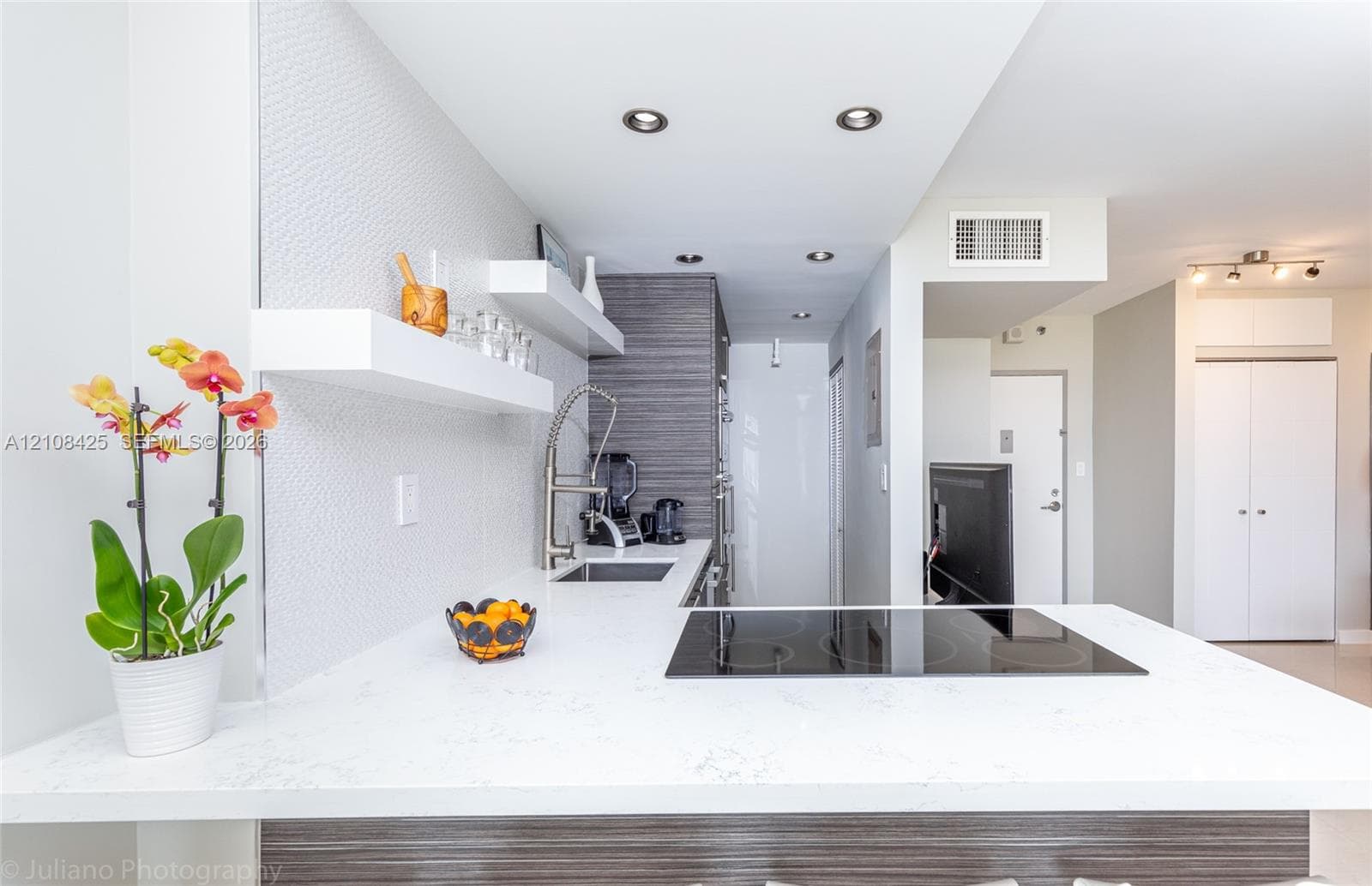 Kitchen featuring a solid-surface countertop with an integrated electric cooktop, floating wall shelving, high-contrast cabinetry, a stainless steel gooseneck faucet, and recessed ceiling lighting