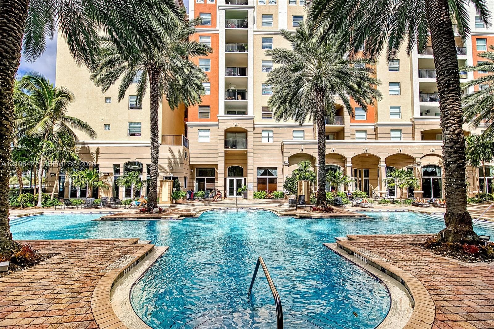Resort-style swimming pool featuring a brick-paver deck, arched architectural detailing, mature palm trees, and multi-story residential facade