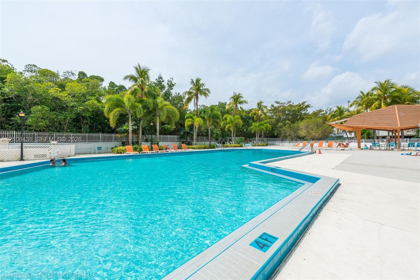 In-ground swimming pool with a tiled perimeter, expansive concrete deck, covered pavilion, and mature perimeter landscaping