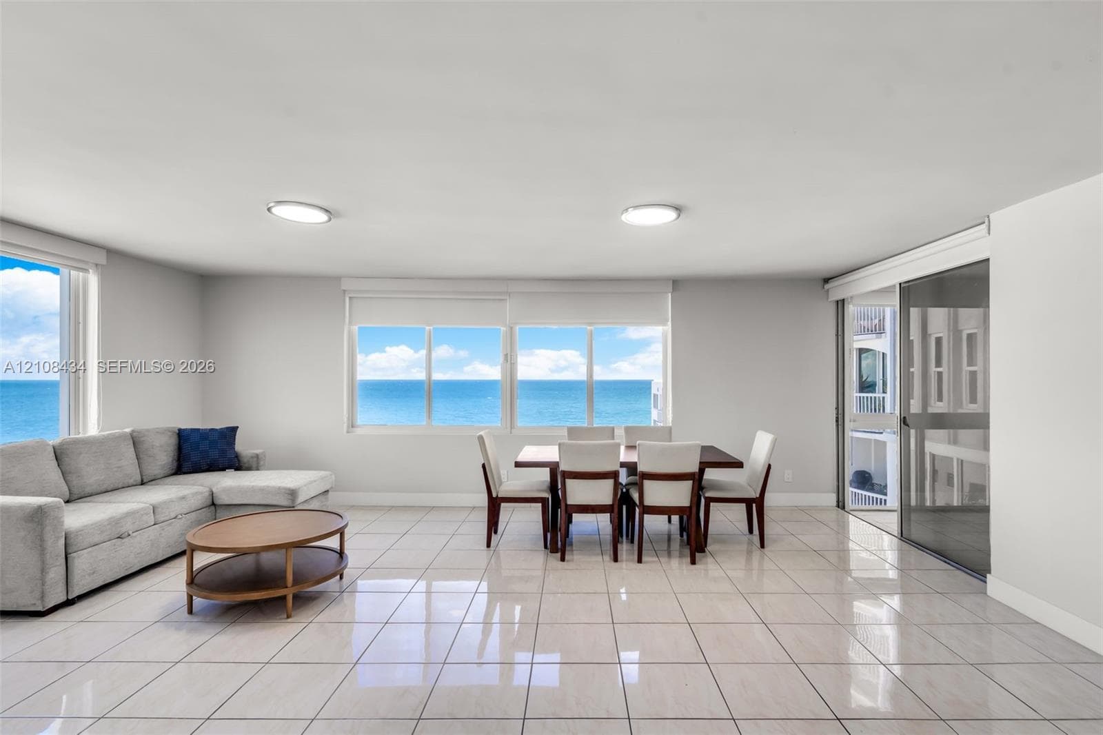 Open-concept room featuring light-toned tile flooring, large multi-pane windows, and a sliding glass door providing access to a balcony with an ocean backdrop