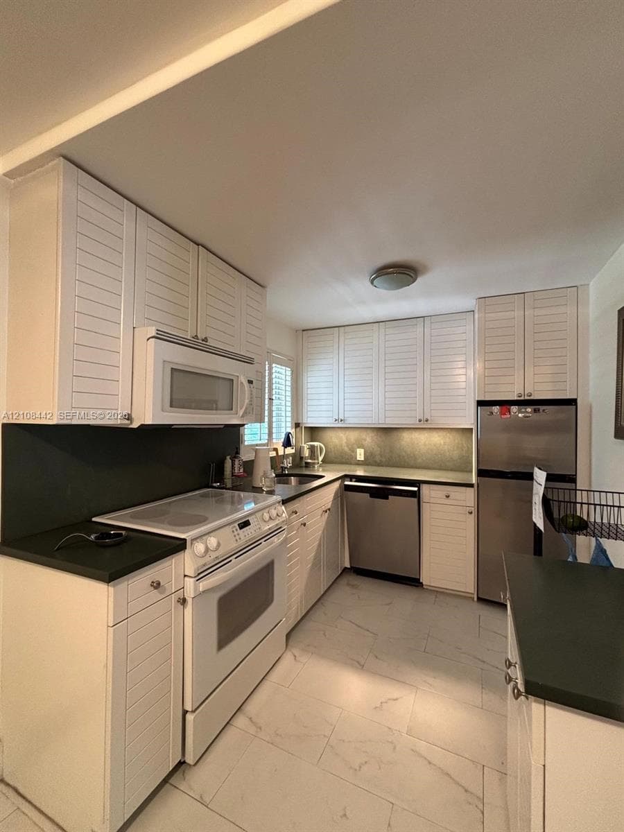 Kitchen featuring white louvered cabinetry, solid surface countertops, light-toned stone-finish flooring, and built-in stainless steel appliances