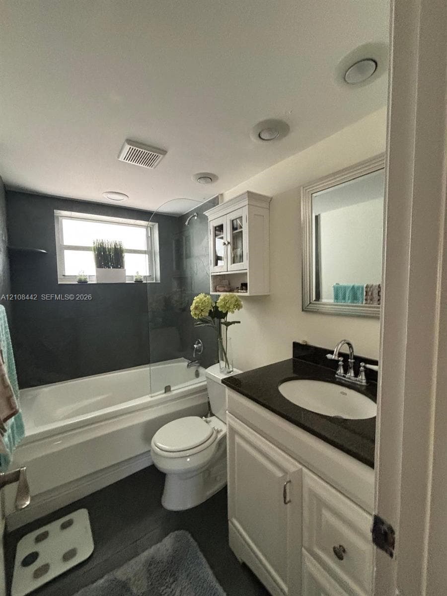 Bathroom featuring a vanity with a dark countertop and integrated sink, white cabinetry, a wall-mounted medicine cabinet, a tub-shower combination with a glass splash panel, and recessed lighting