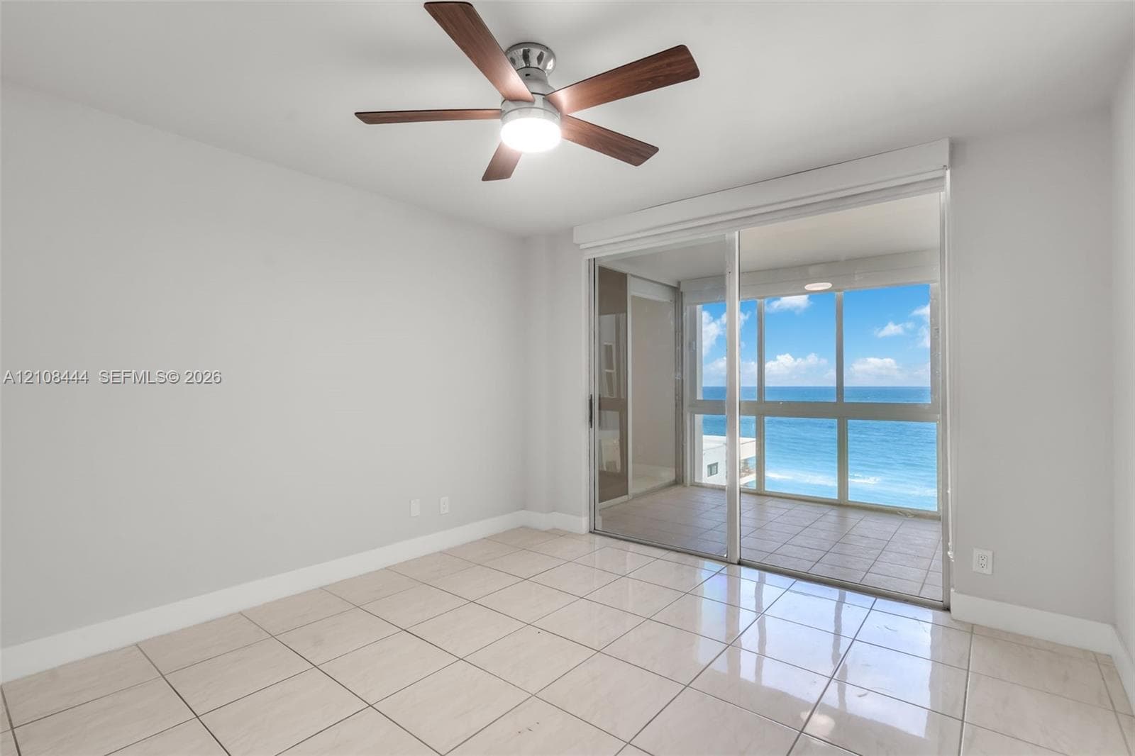 Interior space featuring large-format ceramic tile flooring, ceiling fan with integrated lighting, and full-height sliding glass doors opening to a tiled exterior balcony with an ocean backdrop