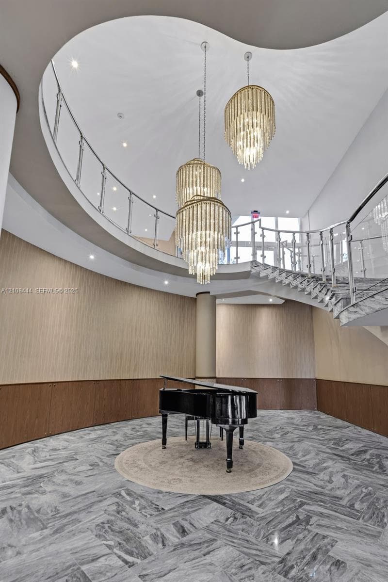 Grand foyer featuring a sweeping curved staircase with polished metal railings, tiered crystal chandeliers, stone flooring in a herringbone pattern, and wood-paneled walls