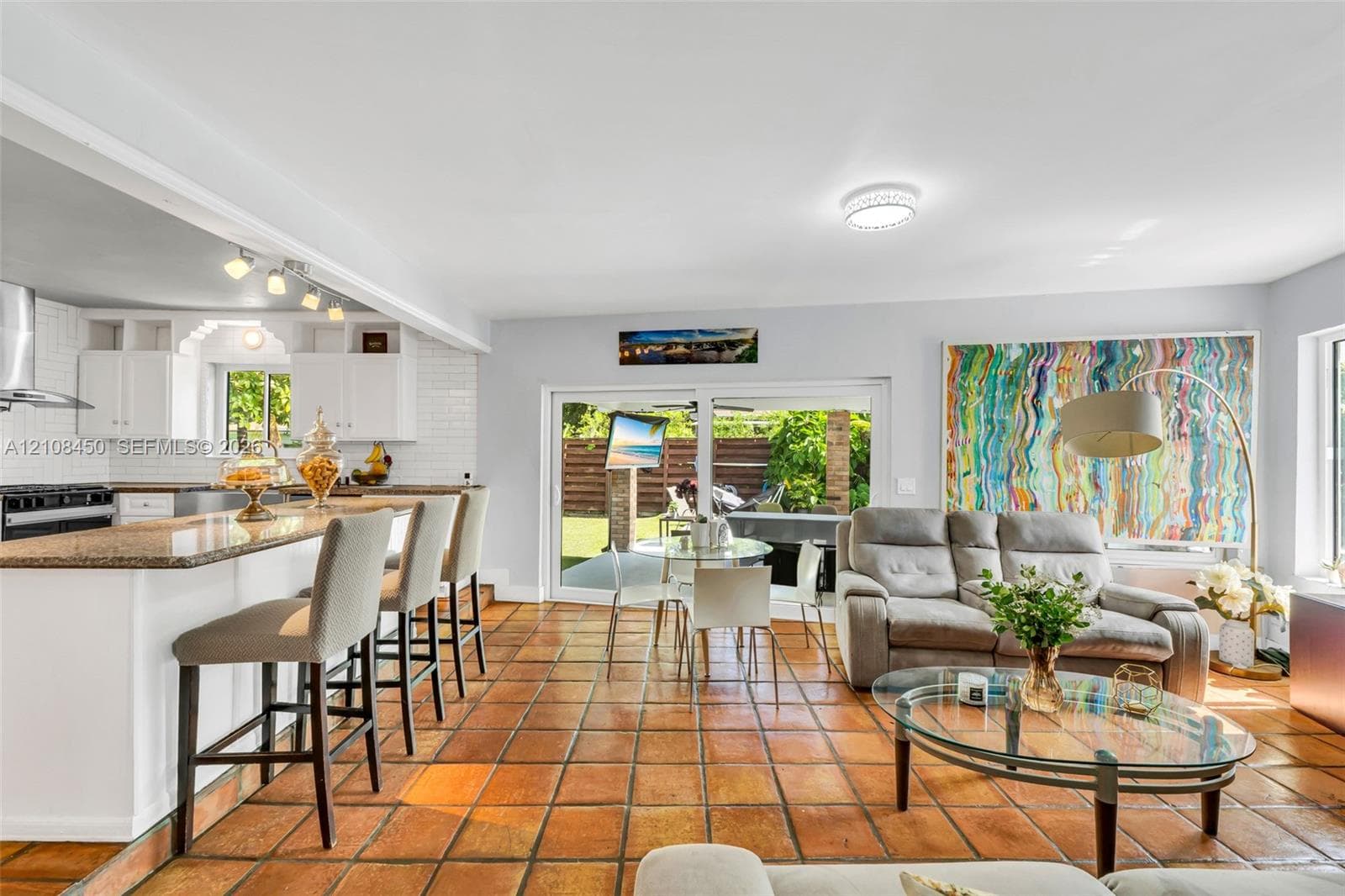Open-concept kitchen and living area featuring ceramic tile flooring, kitchen island with stone countertop, white shaker-style cabinetry, white tile backsplash, and sliding glass doors
