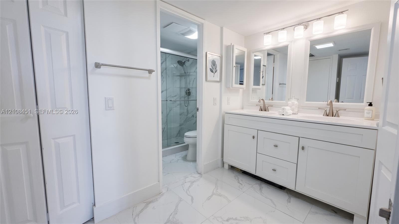 Dual-sink vanity with white cabinetry, light-toned countertop, framed mirrors, glass-enclosed shower with tiled surround, and large-format tile flooring