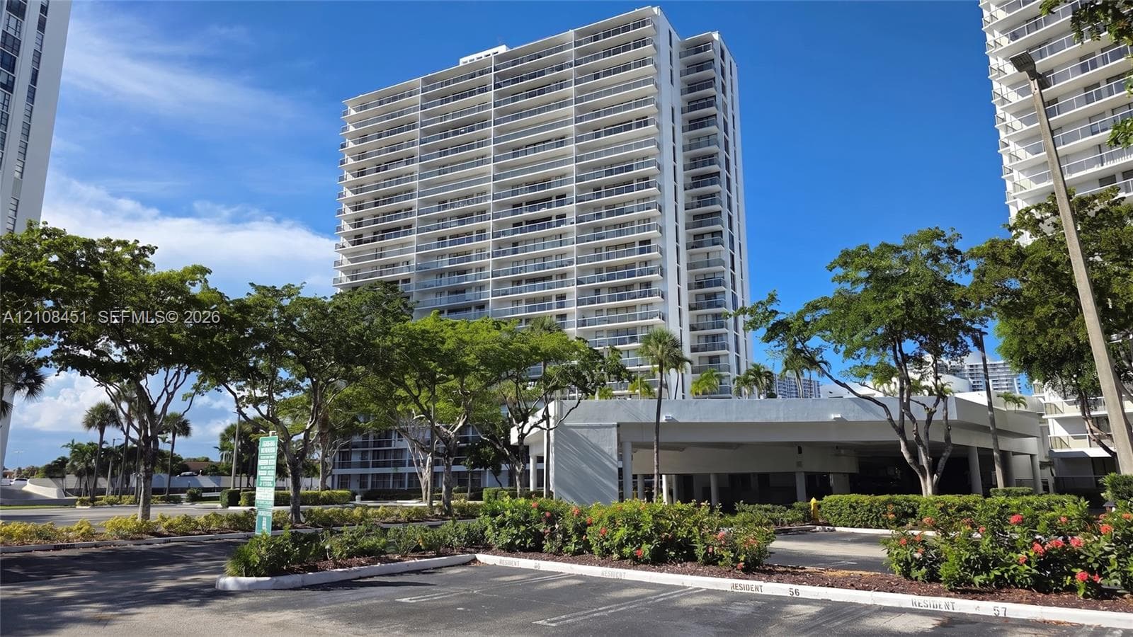 High-rise residential building featuring expansive private balconies, a multi-level concrete structure, and ground-level covered parking, complemented by mature landscaping