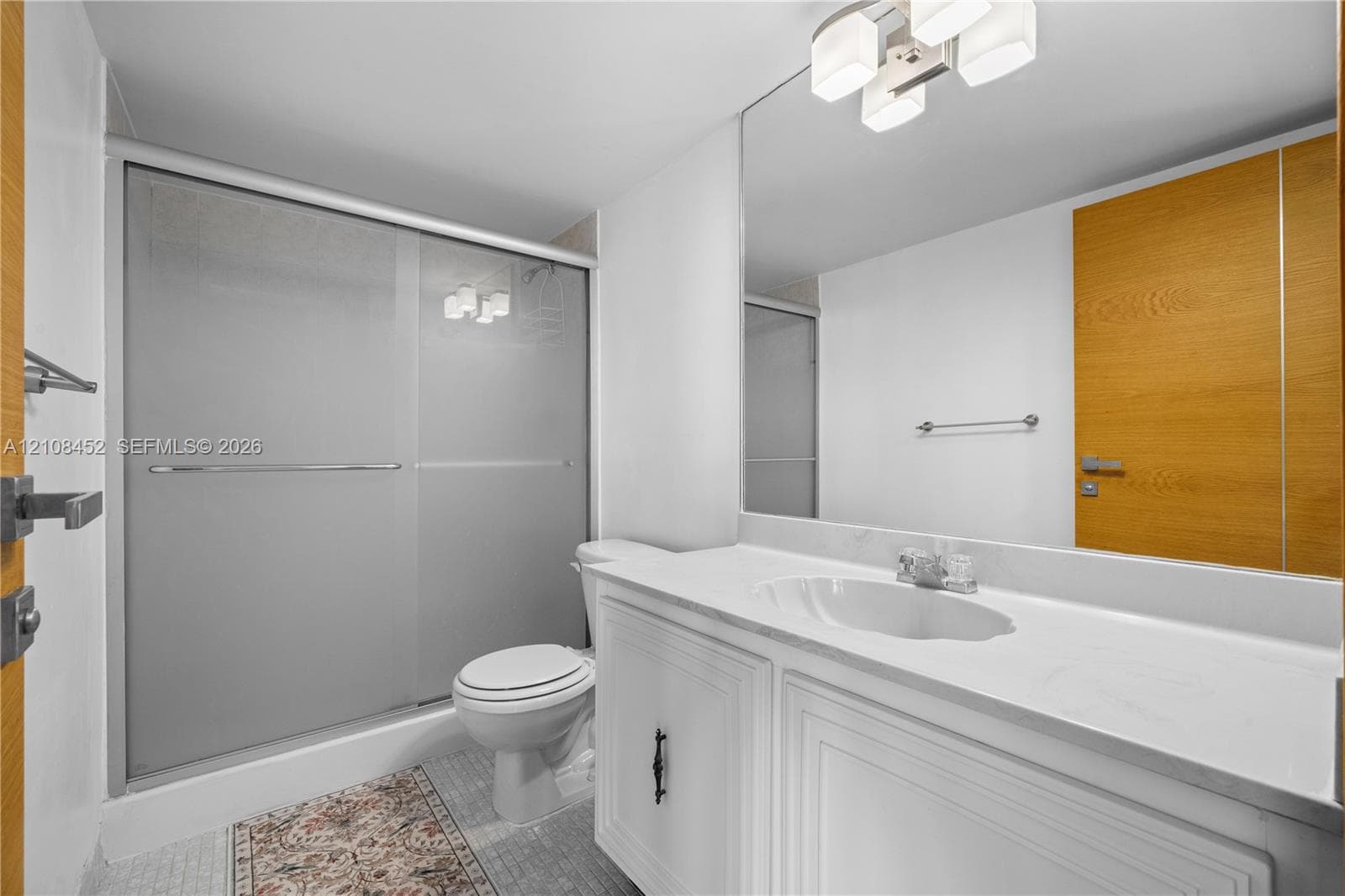 White vanity with a stone countertop, integrated sink, large wall-mounted mirror, sliding glass shower enclosure, and modern multi-light fixture