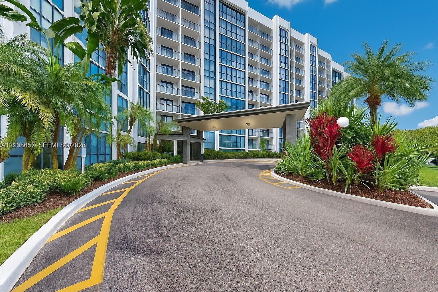 High-rise building exterior featuring a covered porte-cochère, glass facade, expansive balcony tiers, and landscaped grounds with mature palm trees