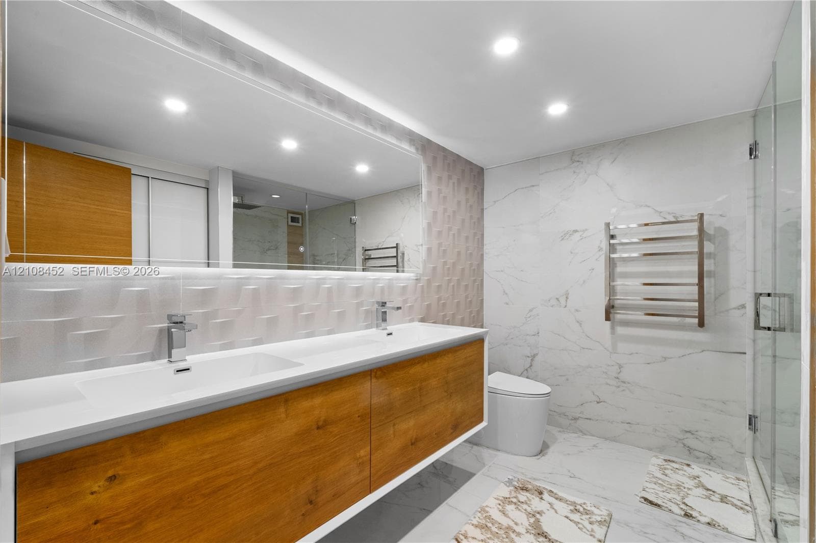 Floating wood-finish double vanity with rectangular integrated sinks, full-width mirror, recessed ceiling lighting, glass-enclosed shower, and heated towel rack