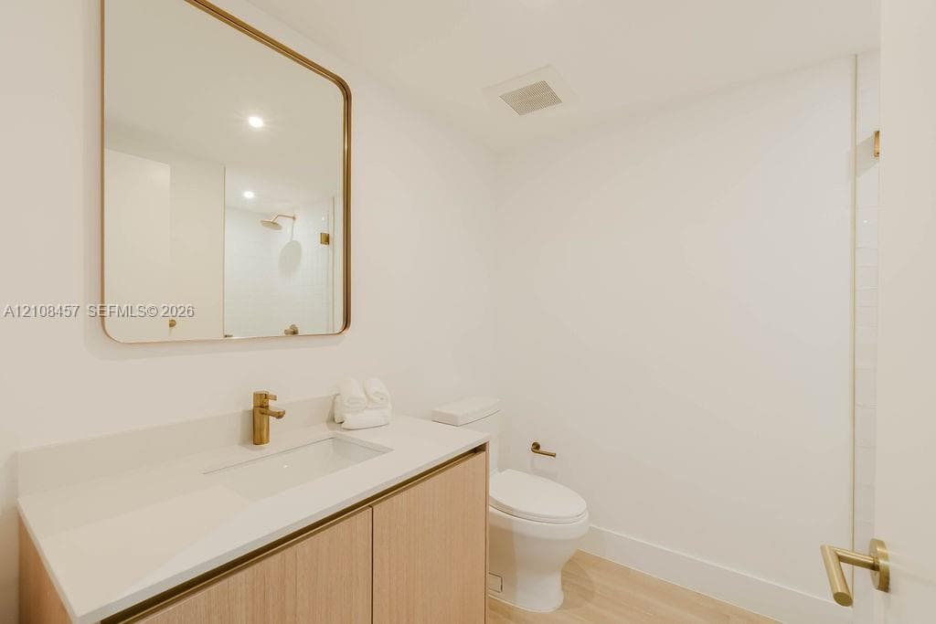 Modern vanity with wood-finish cabinetry, white countertop, integrated rectangular sink, brass-toned hardware, and a matching rounded-edge wall mirror